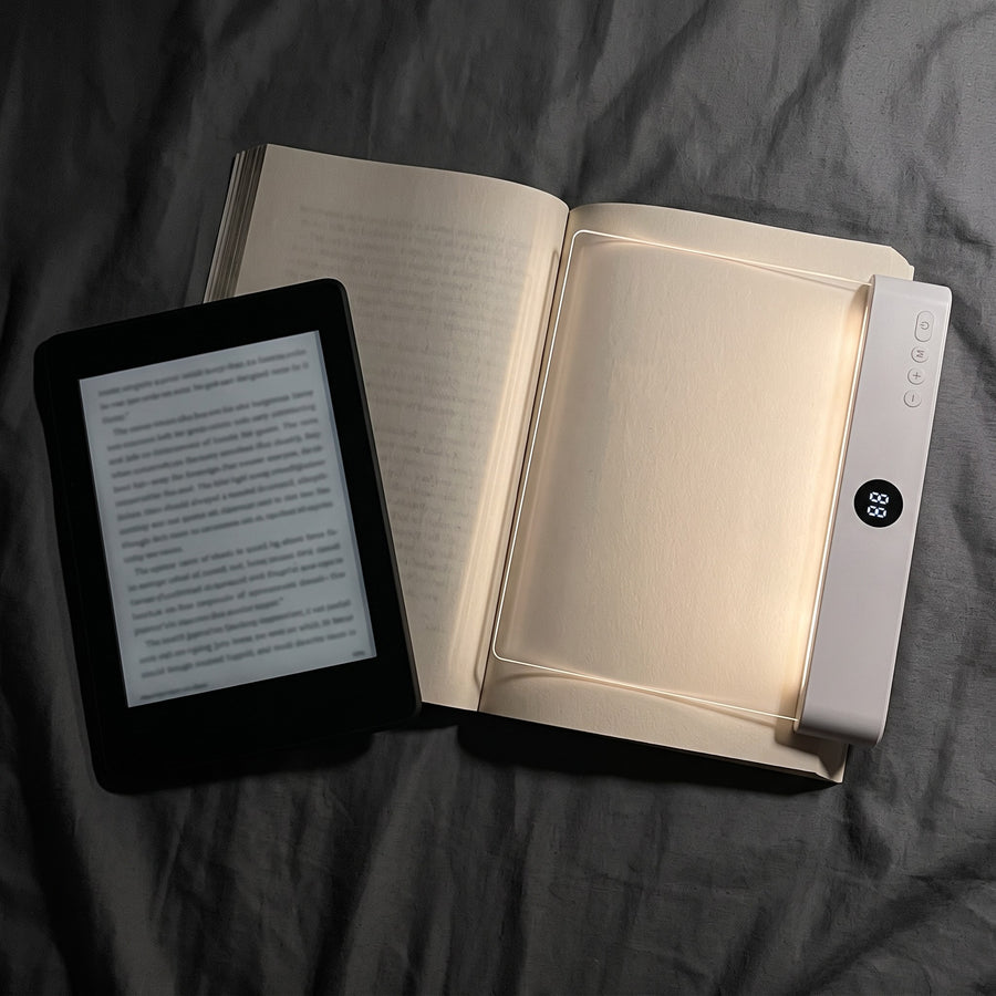 The reading light on a paperback page next to an e-reader, both glowing at night